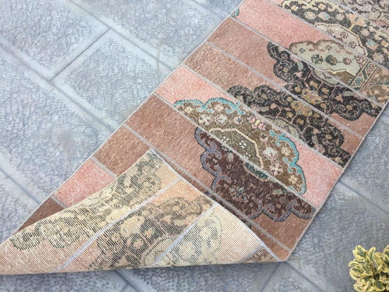 May include: A vintage runner rug with a pink and brown color scheme. The rug has a geometric pattern with a floral design. The rug is folded in half, showing the back side of the rug.