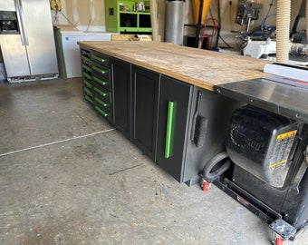 May include: A black and green workbench with a wooden top and drawers. The workbench is in a garage and has a table saw on the right side.