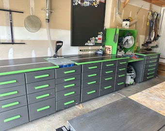 May include: A black and green workbench with multiple drawers. The workbench has a black countertop and a green trim around the edge. There is a saw on the workbench and a vacuum cleaner on the floor.