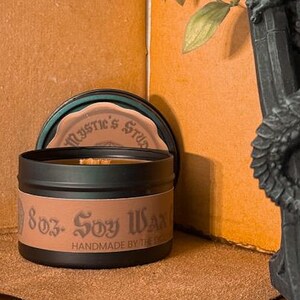 May include: A black soy wax candle in a tin with the text "8oz - Soy Wax" and "Handmade by the Dead". The candle sits on a brown surface, next to a decorative gargoyle statue and green leafy vines.