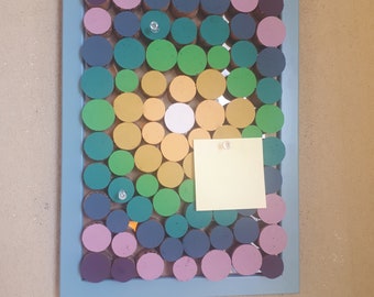 May include: A light blue framed corkboard with a rainbow pattern of painted cork circles. A yellow sticky note is attached to the board with a push pin.