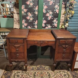 May include: A vintage wooden desk with a dark brown finish. The desk features two side cabinets with drawers and decorative pulls, and a central kneehole area. The desk is set on ornate legs and sits on a patterned rug.