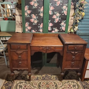 May include: A vintage wooden desk with a dark brown finish. The desk has two side cabinets with drawers and decorative pulls, and a central area with a carved design. The desk is set on a patterned rug.