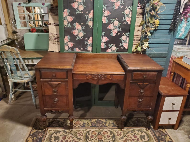 May include: A vintage wooden desk with a dark brown finish. The desk has two side cabinets with drawers and decorative pulls, and a central area with a carved design. The desk is set on a patterned rug.