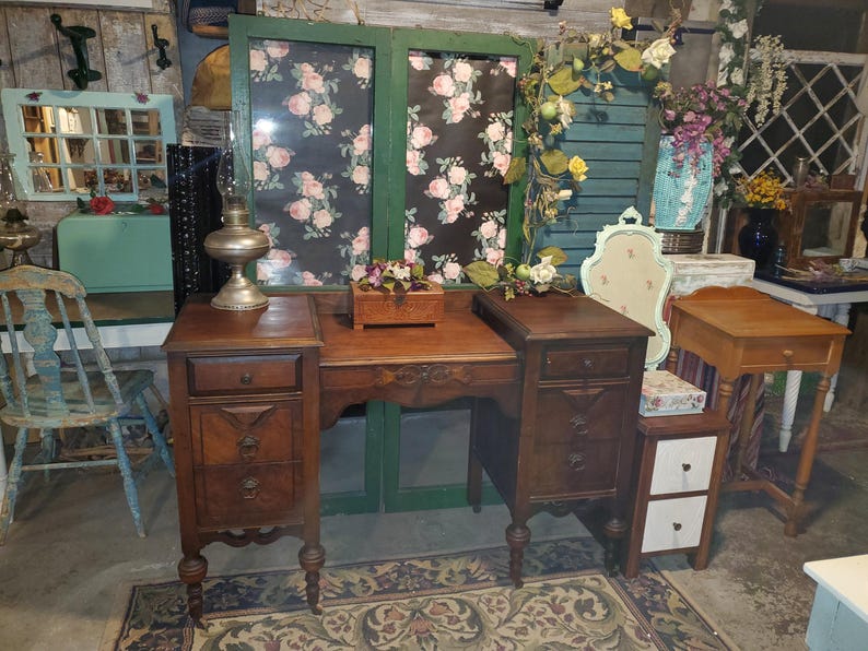 May include: A vintage wooden vanity with two side cabinets, each with drawers and ornate detailing. The vanity is brown, with a floral patterned backdrop and decorative elements, suggesting a classic, antique style.