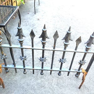 May include: A black metal fence section featuring vertical bars topped with pointed spearhead finials and decorative star-shaped accents. The structure includes two horizontal rails and shows signs of rust at the base and along the metal frame.