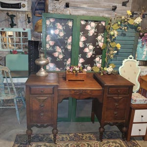 May include: A vintage wooden desk with three drawers on each side, featuring ornate carvings and a warm brown finish. The desk is set on a patterned rug, with a decorative lamp and small wooden box on top. The background includes floral-patterned panels.