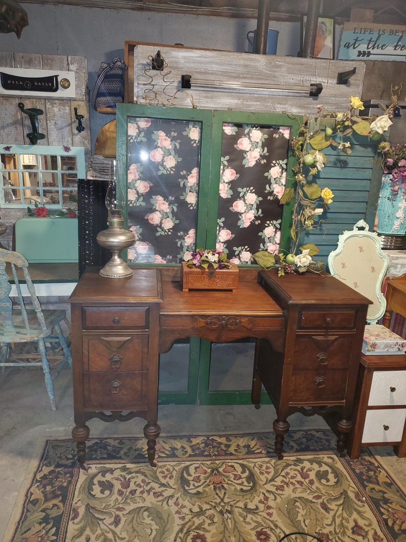 May include: A vintage wooden desk with three drawers on each side, featuring ornate carvings and a warm brown finish. The desk is set on a patterned rug, with a decorative lamp and small wooden box on top. The background includes floral-patterned panels.