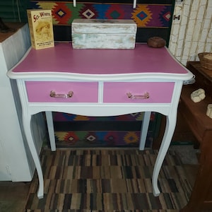 May include: A white and pink vanity table with two drawers and a scalloped edge. The table has white legs with a curved design. The drawers have clear glass handles.