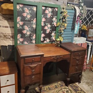 May include: A vintage wooden vanity with a floral patterned backdrop. The vanity has a central desk area with drawers on either side. A small side table with two white drawers is on the left. The vanity is set on a patterned rug.