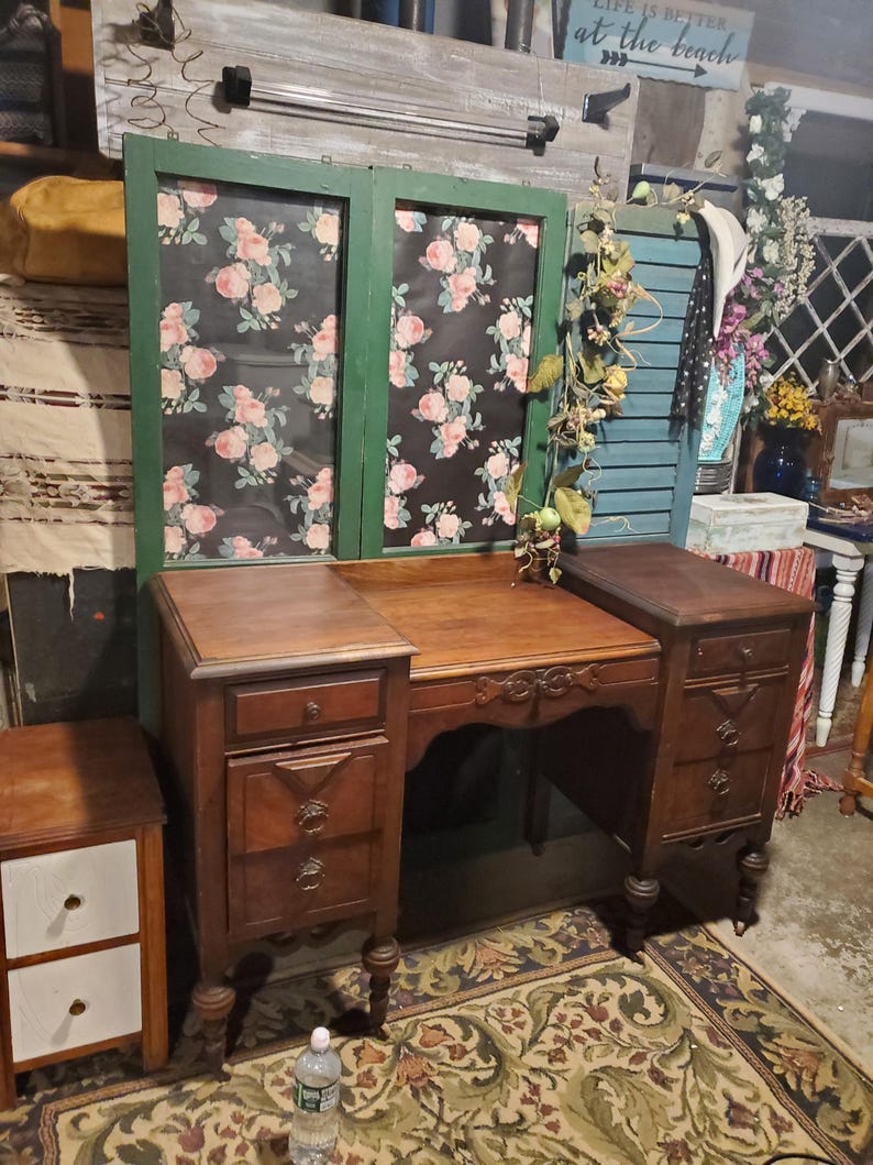 May include: A vintage wooden vanity with a floral patterned backdrop. The vanity has a central desk area with drawers on either side. A small side table with two white drawers is on the left. The vanity is set on a patterned rug.
