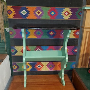 May include: A small, decorative table with a dark brown top and a light green base. The table has a shelf and curved legs. The background features a colorful, patterned textile with geometric designs.