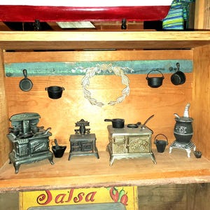 May include: A wooden display case featuring miniature cast iron kitchen items. The display includes small stoves, pots, and pans, with a decorative wreath and painted accents. The overall theme is rustic and vintage.