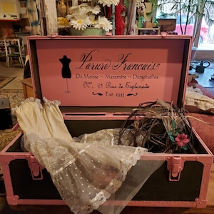May include: A vintage pink and black trunk with the text "Parure Francais" and other French text. Inside the trunk is a cream-colored dress with lace and polka dots, and a decorative wreath. The trunk has pink metal accents.