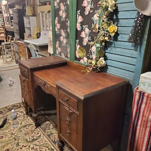 May include: A vintage wooden desk with multiple drawers and ornate detailing. The desk is a rich brown color and features a curved front. The desk is set on a patterned rug. A floral garland decorates a window in the background.