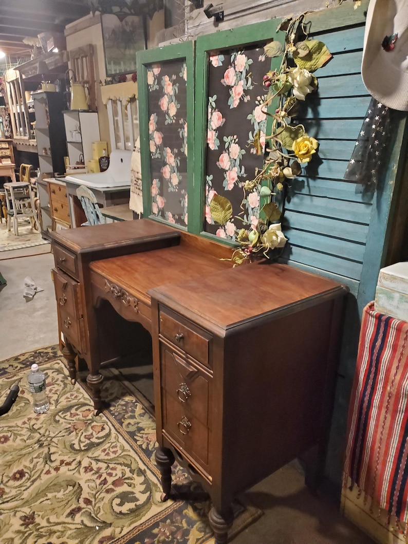 May include: A vintage wooden desk with multiple drawers and ornate detailing. The desk is a rich brown color and features a curved front. The desk is set on a patterned rug. A floral garland decorates a window in the background.