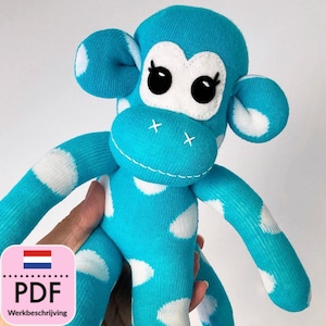 Op de afbeelding: Een turquoise blauwe en witte stippen sok aap met zwarte ogen en een witte neus. De aap heeft een gestikte X op zijn neus en houdt een PDF-patroon vast om een sok aap te maken.