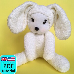 May include: A handmade white bunny doll with long ears, black bead eyes, and a black nose. The bunny has pink cheeks and is sitting with its legs spread. The doll is made from soft, fluffy material. The image includes a "PDF tutorial" graphic.