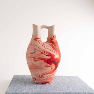 May include: A red and white ceramic vase with a marbled design. The vase has a unique double-spout design and a handle.