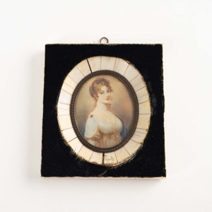 May include: A framed miniature portrait of a woman with auburn hair wearing a white dress and a blue shawl. The frame is made of ivory and has a black velvet backing.