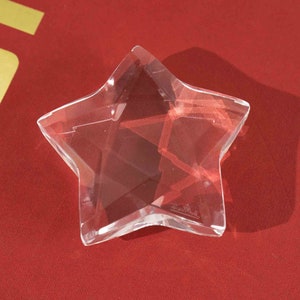 May include: A clear glass star-shaped paperweight with a faceted design. The star is sitting on a red surface.