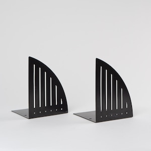 Puede incluir: Un par de sujetalibros de metal negro con forma de semicírculo con líneas verticales recortadas. Los sujetalibros están inclinados para sostener los libros en posición vertical.
