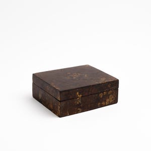 May include: A rectangular lidded box featuring a dark brown surface with a mottled, marbled pattern in shades of tan and gold. The box has a smooth, flat top and straight sides, with a visible seam where the lid meets the base of the container.