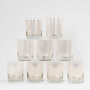 May include: A set of ten clear drinking glasses with vertical gold and silver stripes. The glasses are cylindrical with a thick base and a small logo at the bottom. The glasses are arranged in a pyramid shape.