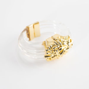 Puede incluir: Un brazalete de plástico transparente con un diseño floral adornado en tono dorado. El brazalete está hecho de múltiples capas de plástico transparente.