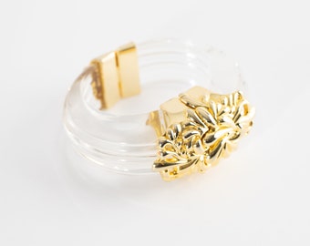 May include: A clear plastic bangle bracelet with a gold-toned, ornate floral design. The bracelet is made of multiple layers of clear plastic.