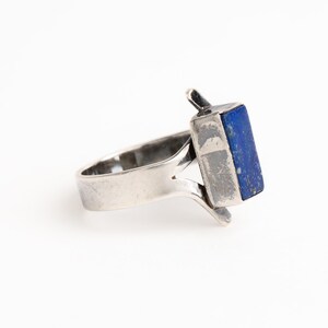 May include: A silver ring with a blue lapis lazuli stone set in a rectangular shape. The ring has a unique design with a split band and a raised setting for the stone.