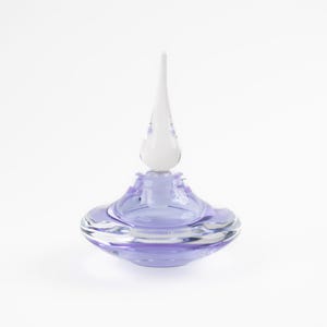 May include: A decorative glass perfume bottle with a light purple base and a white, teardrop-shaped stopper. The bottle has a rounded, saucer-like shape with a clear, wavy rim. The stopper is elongated and tapers to a point.