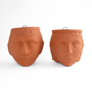 May include: Two terracotta planters shaped like human faces. The planter on the left is a man's face with a beard, and the planter on the right is a woman's face.