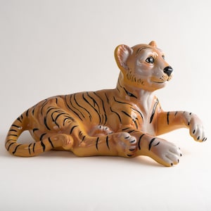 May include: A ceramic tiger figurine in a lying down position. The tiger is orange and black with a white chest and belly. The figurine is detailed with black stripes and a white face.