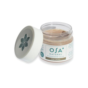 May include: An open jar of Osa Natural sunscreen with the lid beside it. The jar is clear, revealing a light brown cream. The lid is white with a green floral design. Text on the jar reads "Bloqueador Solar Con Cacao".