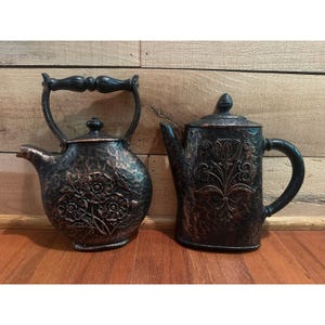 May include: Two dark metal teapots with a hammered texture and copper-toned highlights. The round teapot features a floral relief and a swing handle, while the taller, cylindrical teapot displays a vertical floral motif and a side handle.