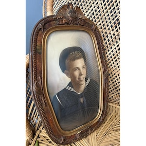 May include: An oval-shaped wooden picture frame featuring a carved eagle at the top and nautical motifs along the border. The frame contains a vintage portrait of a service member wearing a dark navy uniform and a cap with the text U.S. NAVY.