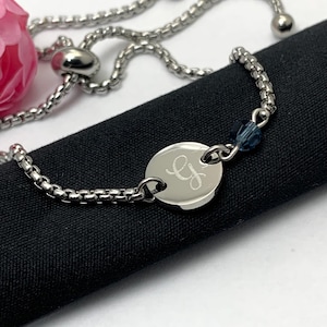 May include: A silver bracelet with a box chain design. The bracelet has a circular charm engraved with the initials "ey" and a small blue bead. The bracelet is displayed on a black surface.