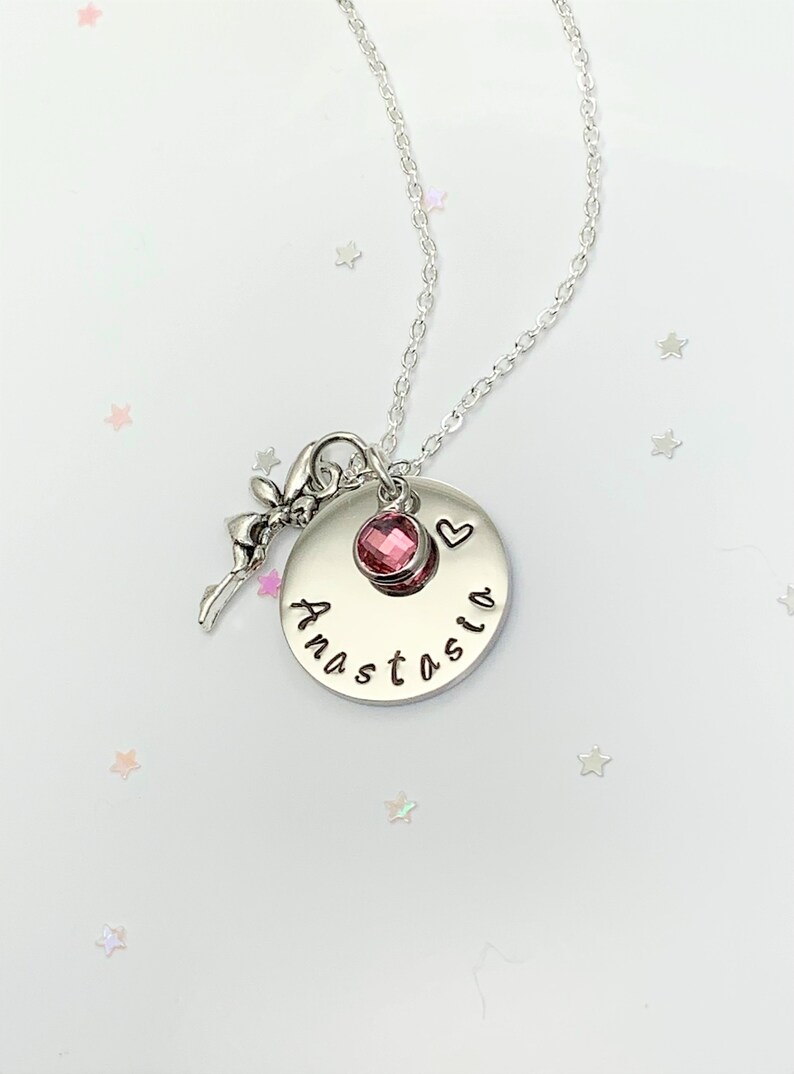 K&ouml;nnte beinhalten: Eine silberne Halskette mit einem Anh&auml;nger, auf dem "Anastasia" steht und der einen kleinen rosa Kristall und einen Feenanh&auml;nger hat.