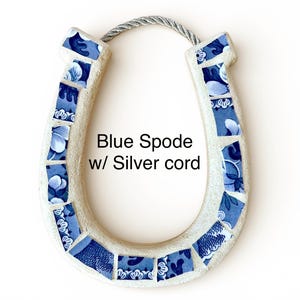 May include: A horseshoe-shaped wall hanging with a mosaic design. The horseshoe is cream-colored with blue and white floral patterned tiles. It has a silver cord for hanging. The text "Blue Spode w/ Silver cord" is visible.