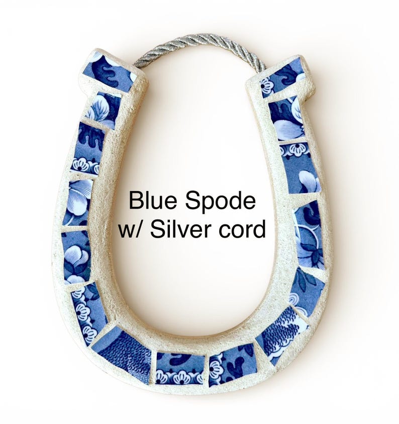 May include: A horseshoe-shaped wall hanging with a mosaic design. The horseshoe is cream-colored with blue and white floral patterned tiles. It has a silver cord for hanging. The text "Blue Spode w/ Silver cord" is visible.