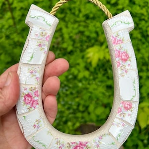 May include: A mosaic horseshoe decoration made from white ceramic pieces with pink rose patterns. The horseshoe is accented with a gold rope for hanging. The background is a blurred green.
