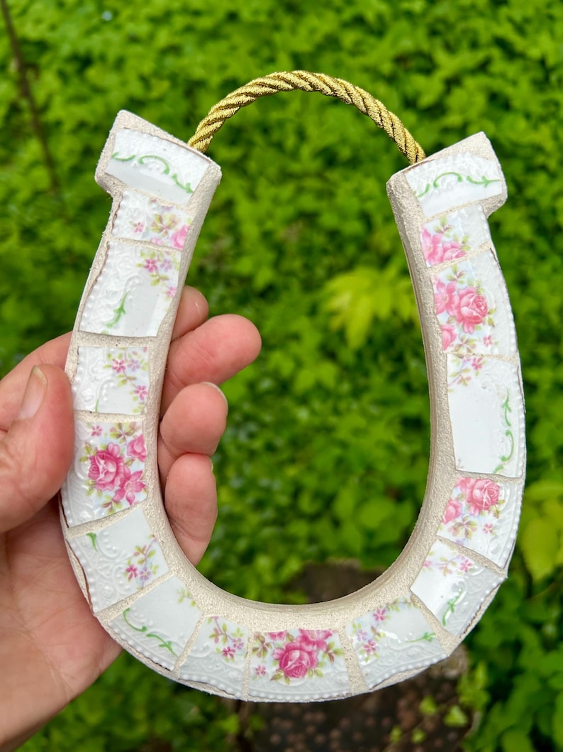 May include: A mosaic horseshoe decoration made from white ceramic pieces with pink rose patterns. The horseshoe is accented with a gold rope for hanging. The background is a blurred green.