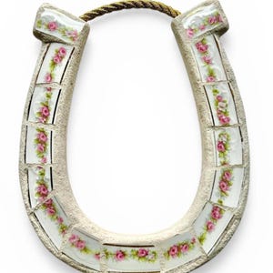 May include: A decorative horseshoe with a mosaic design. The horseshoe is made of a light beige material and is adorned with rectangular tiles featuring pink roses and green leaves. A gold-colored rope is attached at the top.