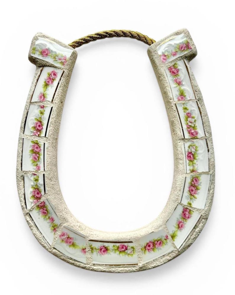 May include: A decorative horseshoe with a mosaic design. The horseshoe is made of a light beige material and is adorned with rectangular tiles featuring pink roses and green leaves. A gold-colored rope is attached at the top.