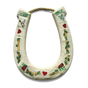 May include: A cream-colored horseshoe-shaped wall hanging with mosaic tile accents. The tiles feature images of hearts, birds, and floral patterns. A gold rope is attached for hanging. The horseshoe is a symbol of good luck.