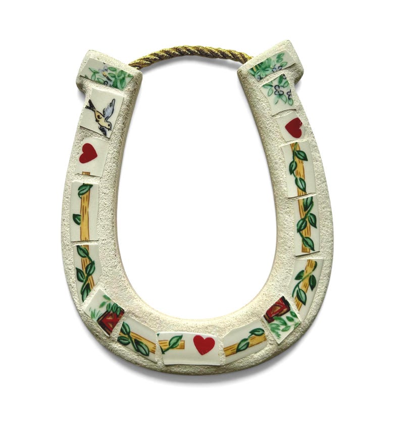 May include: A cream-colored horseshoe-shaped wall hanging with mosaic tile accents. The tiles feature images of hearts, birds, and floral patterns. A gold rope is attached for hanging. The horseshoe is a symbol of good luck.