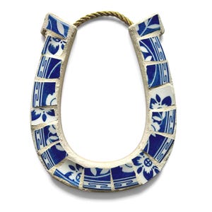May include: A horseshoe-shaped decorative item made from broken blue and white ceramic tiles. The tiles are arranged in a mosaic pattern and are connected by a light-colored grout. A gold-colored rope is attached at the top.