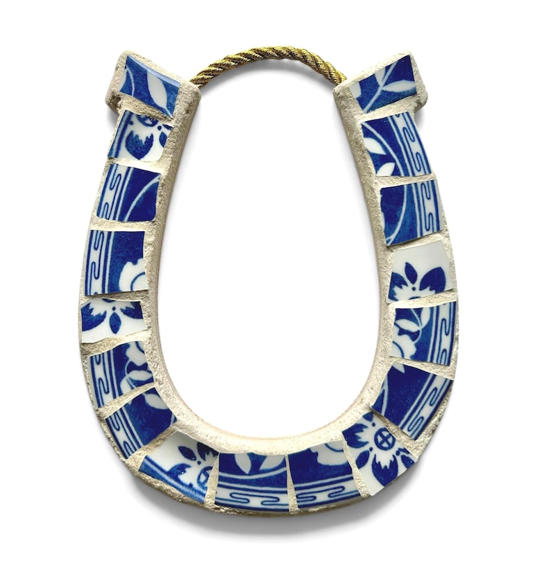 May include: A horseshoe-shaped decorative item made from broken blue and white ceramic tiles. The tiles are arranged in a mosaic pattern and are connected by a light-colored grout. A gold-colored rope is attached at the top.