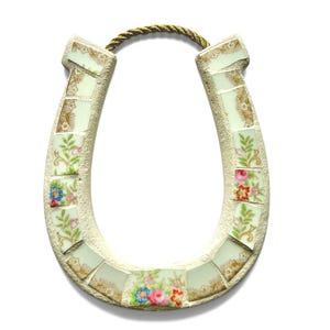 May include: A decorative horseshoe made of mosaic tiles with floral patterns and gold accents. The horseshoe is cream-colored with a gold rope for hanging. The mosaic tiles feature pink, blue, and red flowers.